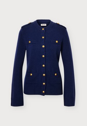 Femme aux cheveux foncés portant un cardigan bleu marine boutonné et un pantalon beige ample, debout devant un fond clair uni.