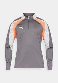 Šedé dlouhé sportovní tričko s polovičním zipem, s oranžovými a šedými barevnými bloky, které má bílé logo na hrudi.