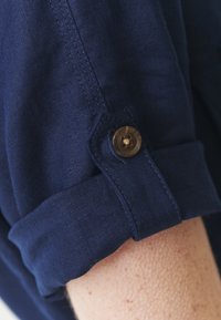 Košile s rukávy ohrnutými do námořnické modré barvy, vybavená knoflíkovým zapínáním, vyrobená z texturované látky s viditelnými prošitými detaily.