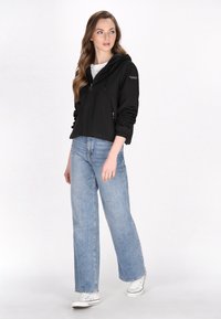 Jachetă neagră cu glugă, cu fermoar în față, manșete elastice la mâneci și două buzunare frontale, purtată cu blugi evazați de culoare albastră deschisă și adidași albi.