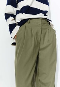 Pantalons plissés vert olive à taille haute, associés à une chemise à rayures bleues et blanches à manches longues, présentés sur un fond clair.