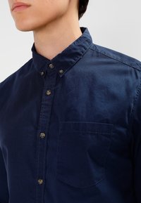 Tummansininen nappikauluspaita, jossa on teksturoitu kangas, rintatasku ja tummat napit. Tasainen pinta, ei näkyviä kuvioita.