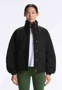 Person iført en sort quiltet jakke over en hvid skjorte og grønne bukser, vendt fremad med et neutralt udtryk mod en ensfarvet baggrund.