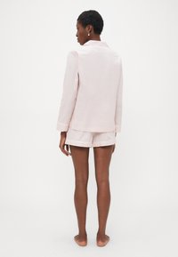 Kvinne som står barfot, med ryggen til, iført en lys rosa langermet pysjamasoverdel og matchende shorts mot en ensfarget bakgrunn.