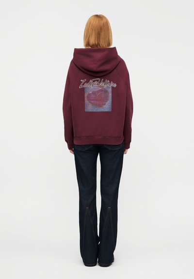Maroon hoodie with a large heart graphic on the back and "Zadig&Voltaire" text. Paired with dark blue flared jeans. Soft material.