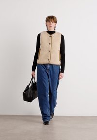 Μπεζ γούνινο γιλέκο με υφή, καφέ πλαϊνές τσέπες και κουμπότρυπα μπροστά. Φοριέται πάνω από έναν μαύρο ζιβάγκο, σε συνδυασμό με φαρδιά μπλε τζιν και μια μαύρη τσάντα.