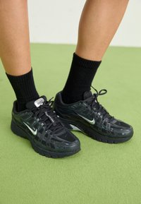 Black Nike sneakers with mesh and synthetic upper, featuring reflective accents, thick soles, and flat laces. Worn with black ribbed socks.