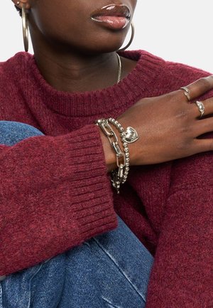 Des bracelets en argent avec des perles et un charm en forme de cœur au poignet, portés avec un pull bordeaux et un jean. Les accessoires ont des accents métalliques.