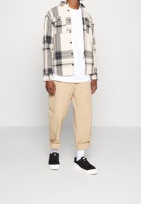 Koszula flanelowa w kratę w kolorach beżowym, szarym i czarnym, w połączeniu z białym t-shirtem i beżowymi podwiniętymi spodniami. Czarne trampki z białymi podeszwami.