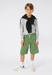 Jongen met kort lichtbruin haar, gekleed in een grijze langsteekshirt, een zwarte sweatshirt rond zijn schouders, groene shorts, witte sokken en sneakers.