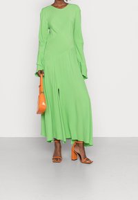 Abito a maniche lunghe di un vivace verde, con una vestibilità leggermente ampia e orlo asimmetrico. Abbinato a sandali arancioni con tacco blocco e una piccola borsa.