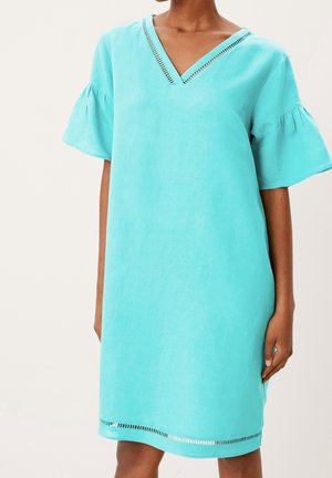 Nainen, joka pukeutunut väljään turkoosiin polvipituiseen mekkoon, jossa on röyhelöhihat ja leikkaukset pääntiessä sekä helman kohdalla, yksiväristä taustaa vasten.