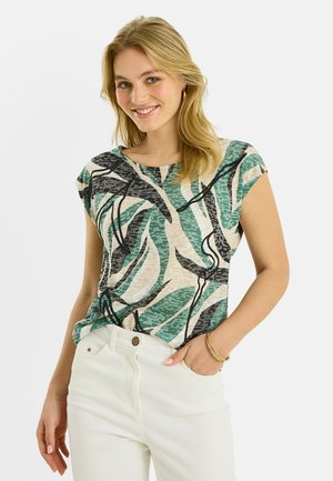 Mujer sonriente con una blusa de manga corta con patrón abstracto en verde, negro y beige, pantalones blancos y pulsera dorada, con una mano en el bolsillo.