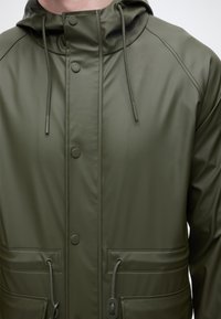 Σκούρο πράσινο αδιάβροχο μπουφάν με κουκούλα, με κουμπιά τύπου snap, ρυθμιζόμενα κορδόνια στην κουκούλα και τσέπες στη μέση, φορεμένο από ένα άτομο.