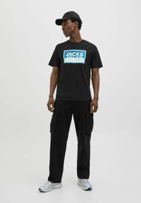 Sort bomulds T-shirt med blåt rektangulært logo med teksten "JACK & JONES." Sammen med sorte cargo-bukser og sølvfarvede sportssko.