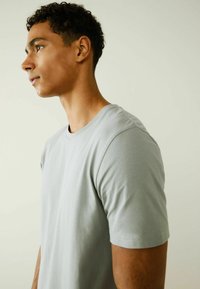 Lichtgrijze katoenen t-shirt met korte mouwen, ronde hals en een gladde textuur, getoond op een model van opzij tegen een neutrale achtergrond.