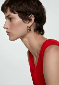 Boucles d'oreilles en or avec un design torsadé, portées par une femme en haut sans manches rouge. Texture lisse, vue de profil contre un fond clair.