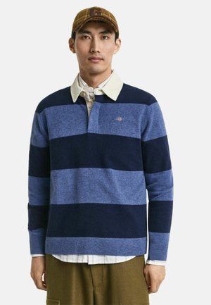 Long-sleeve striped sweater in dark blue and light blue wool blend, featuring a buttoned collar and logo on the chest, worn over a collared shirt.