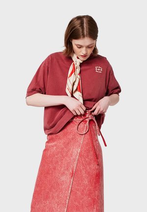 Roestkleurig oversized katoenen t-shirt met korte mouwen en geborduurde accenten, gecombineerd met een rode denim wikkelrok met een strikdetail.
