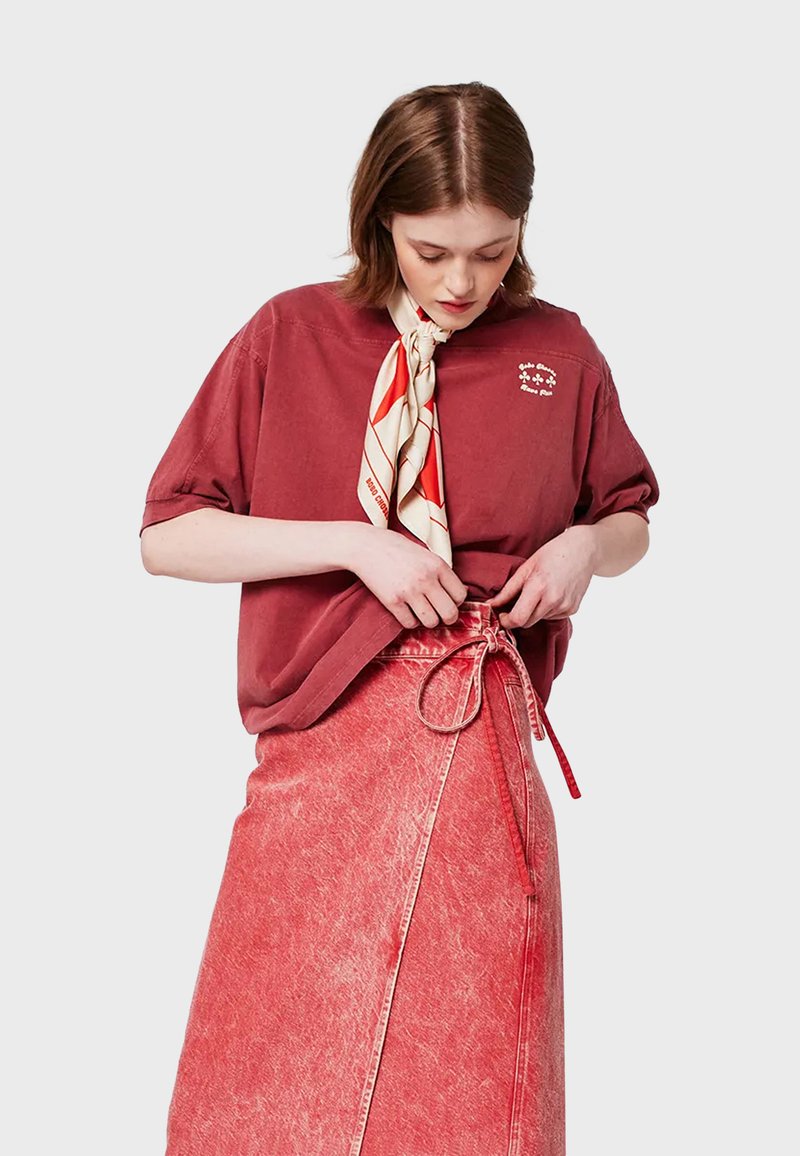 T-shirt in cotone oversize color ruggine con maniche corte e dettagli ricamati, abbinato a una gonna avvolgente in denim rossa con un dettaglio a fiocco.