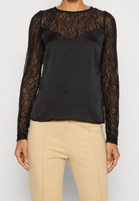 Blouse en satin noir avec manches longues en dentelle florale et poitrine supérieure, associée à un pantalon beige ajusté.