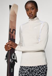 Beige lange mouw rits-top met witte accenten, gecombineerd met zwart-witte patroonbroek en een ski met zwarte bindingen, vastgehouden in de handen.