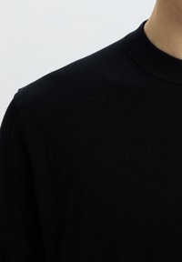 Primo piano di una persona che indossa un maglione nero semplice con scollo tondo su uno sfondo grigio chiaro.