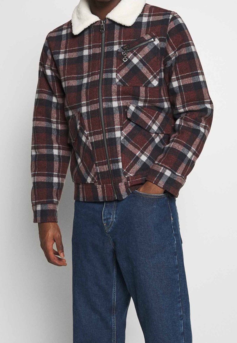 Giacca a quadri rossa e grigia con colletto in pelliccia sintetica bianca, chiusura con zip, tasche multiple e polsini a costine, indossata sopra jeans blu.