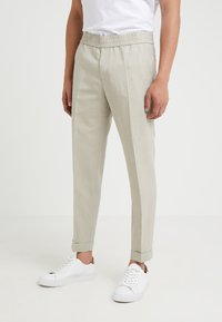 Light beige trousers with a smooth texture, elastic waistband, and side pockets. Rolled cuffs and a tapered fit. Paired with white sneakers.