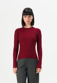 Femme avec des cheveux raides et foncés portant un sweat-shirt noir à manches longues ajusté avec des côtes et un pantalon gris ample devant un fond blanc uni.