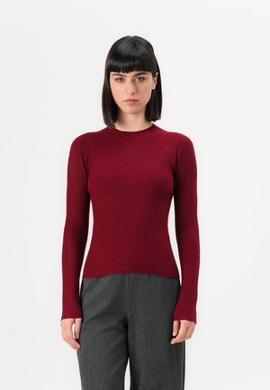 Vrouw met steil donker haar, gekleed in een aansluitende geribbelde donkerrode lange mouw trui en losse grijze broek, tegen een effen witte achtergrond.