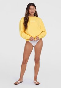Žltá mikina s bielym nápisom "Surf Society", voľný strih, dlhé rukávy, skombinovaná s bielymi bikiny spodkami a žabkami.