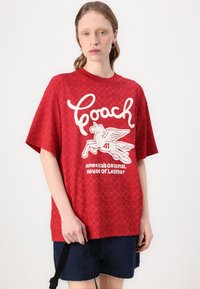T-shirt oversized vermelho com um logo branco "Coach" e um gráfico de cavalo alado, feito de um tecido suave com um padrão texturizado.