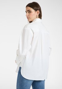 Mujer de cabello oscuro con una camisa blanca holgada abotonada y jeans azules, mirando por encima del hombro contra un fondo liso.