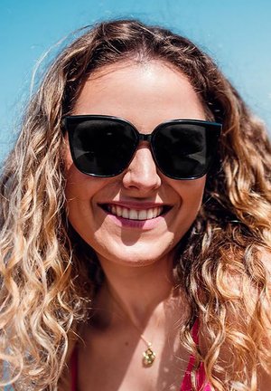 Černé sluneční brýle ve tvaru kočičích očí s lesklým povrchem, které nosí osoba s kudrnatými vlasy. Pozadí tvoří jasná modrá obloha.