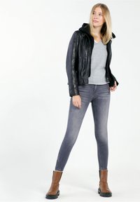 Schwarze Lederjacke mit Kapuze, grauer V-Ausschnitt-Pullover, graue schmale Jeans und braune Stiefeletten mit schwarzen Sohlen. Glatte Oberfläche, tailliertes Design.
