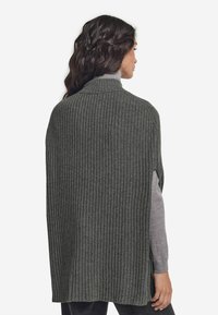 Grå ribbad stickad poncho, öppen på sidorna, hög krage, mjuk textur, sträcker sig ner till mitten av låret, lager på lager över en grå långärmad topp.