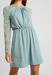 Robe bleu clair avec un corsage plissé et des manches longues en dentelle. Dispose d'une taille élastique, créant une silhouette décontractée. Texture de tissu lisse.