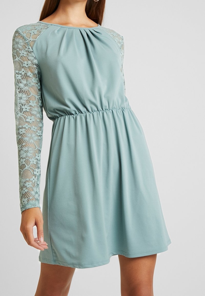 Robe bleu clair avec un corsage plissé et des manches longues en dentelle. Dispose d'une taille élastique, créant une silhouette décontractée. Texture de tissu lisse.