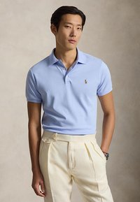 Jeune homme portant un polo bleu clair et un pantalon crème plissé, debout avec une main dans la poche devant un fond uni.