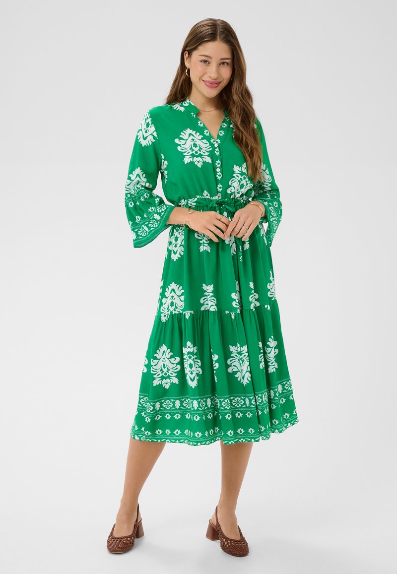 Kobieta stojąca w zielonej sukience midi z białym kwiatowym wzorem, długimi rękawami i brązowymi plecionymi butami na klockowym obcasie, uśmiechająca się z złożonymi dłońmi.