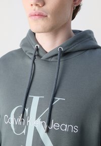 Temnozeleni bombažni hoodie s velikim belim logotipom "CK" in izvezenim besedilom "Calvin Klein Jeans", s sivimi vrvicami in kovinskimi konicami.
