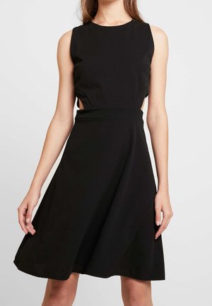 Mouwloze zwarte jurk met zijuitsparingen en een nauwsluitend lijfje dat uitloopt in een A-lijn rok, gedragen door een staande vrouw.