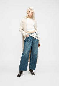 Νεαρή γυναίκα με πλατινένια ξανθά μαλλιά που φοράει μακρύ μπεζ cardigan, λευκό crop top, φαρδιά μπλε τζιν παντελόνια, μαύρα μποτάκια μέχρι τον αστράγαλο και τσάντα μέσης με πλέξη κροσέ.