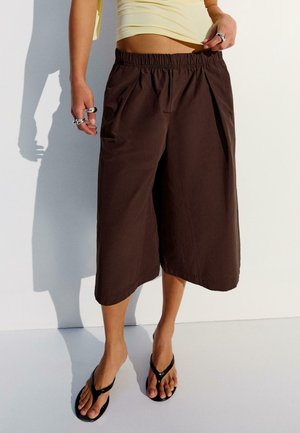 Persoon die losse bruine knielange shorts draagt, een lichtgeel cropped top, zilveren ringen en zwarte teenslippers, staand tegen een witte muur.