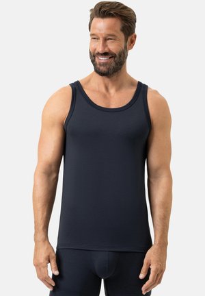 Lächelnder bärtiger Mann, der ein eng anliegendes schwarzes ärmelloses Tanktop und passende schwarze Unterwäsche trägt, steht vor einem einfarbigen hellen Hintergrund.