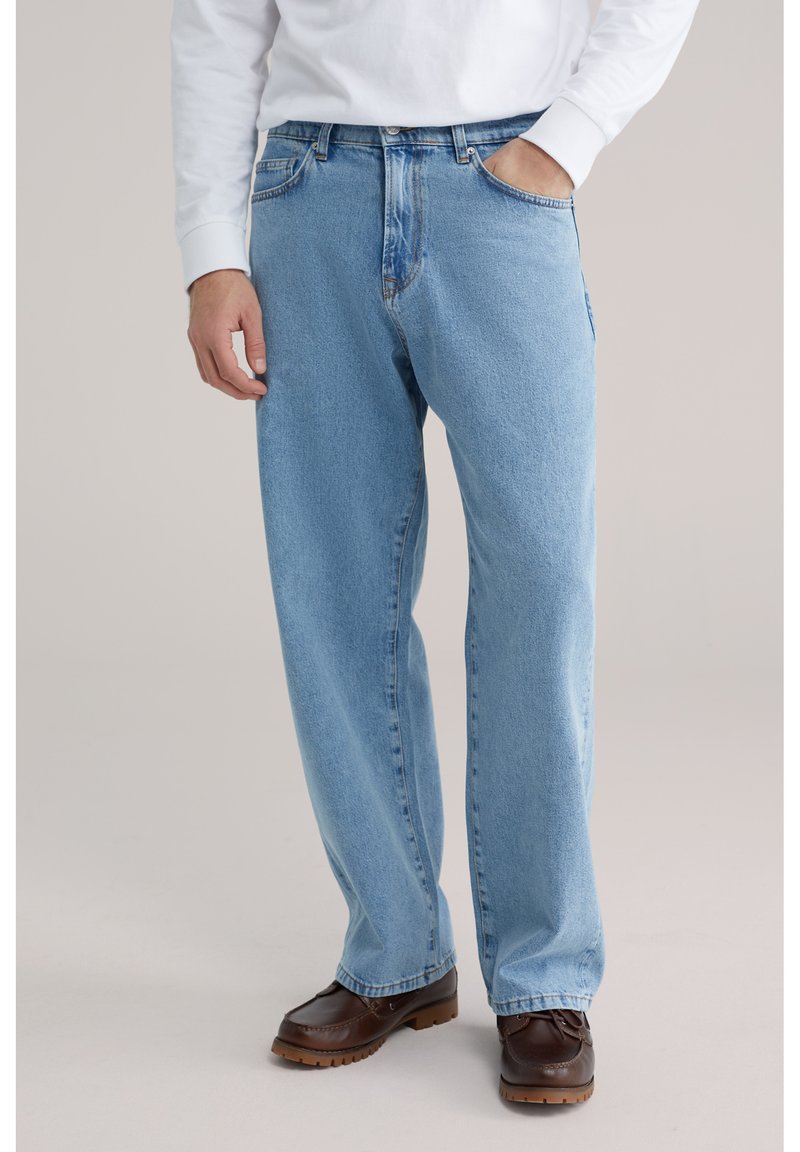 Jean en denim bleu clair à coupe décontractée, doté de cinq poches, passants pour ceinture et d'une finition délavée, accompagné de chaussures marron à enfiler.
