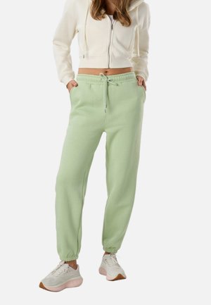 Femme portant un sweat à capuche court crème, pantalon de jogging vert clair avec cordon de serrage, et baskets blanches, debout les mains dans les poches.