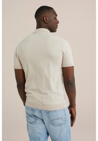 Light beige short-sleeve polo shirt with a ribbed collar and hem, paired with light blue jeans, photographed from the back. Smooth texture.