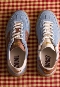 Paire de baskets en daim bleu avec des touches de cuir marron et blanc et des lacets blancs placées sur un tissu à carreaux rouge et jaune.